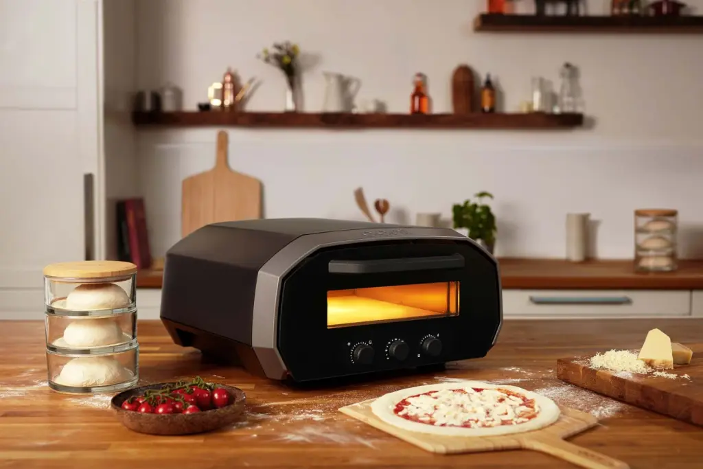 Best indoor pizza oven in a modern kitchen on a wooden countertop. The oven's warm light glows as it preheats. In the foreground, a pizza topped with cheese is ready for baking on a wooden pizza peel. Nearby, a cutting board holds grated cheese and a block of Parmesan. A stack of dough balls in glass containers and a bowl of cherry tomatoes are placed next to the oven. The background features kitchen shelves with various jars, plants, and a wooden pizza paddle.