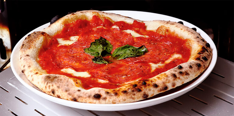 Close-up of a freshly baked Neapolitan-style pizza with a perfectly blistered crust. The pizza is topped with vibrant red tomato sauce, thinly sliced soppressata, melted mozzarella cheese, and fresh basil leaves, served on a white plate. The pizza is sitting on a metal oven rack, highlighting the crispy, airy edges of the crust.