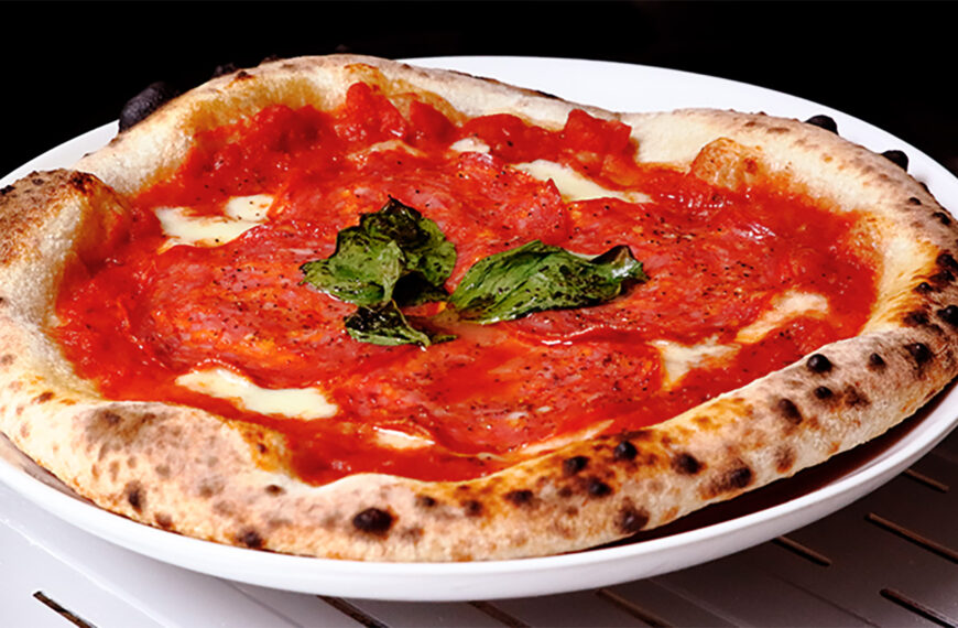 Close-up of a freshly baked Neapolitan-style pizza with a perfectly blistered crust. The pizza is topped with vibrant red tomato sauce, thinly sliced soppressata, melted mozzarella cheese, and fresh basil leaves, served on a white plate. The pizza is sitting on a metal oven rack, highlighting the crispy, airy edges of the crust.