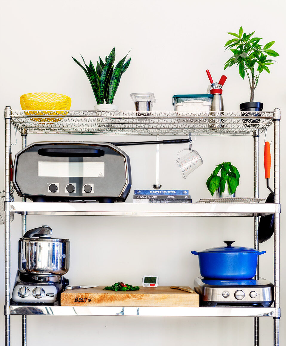 Organized vertical kitchen shelving unit with cooking tools, appliances, and fresh ingredients in a small urban kitchen.